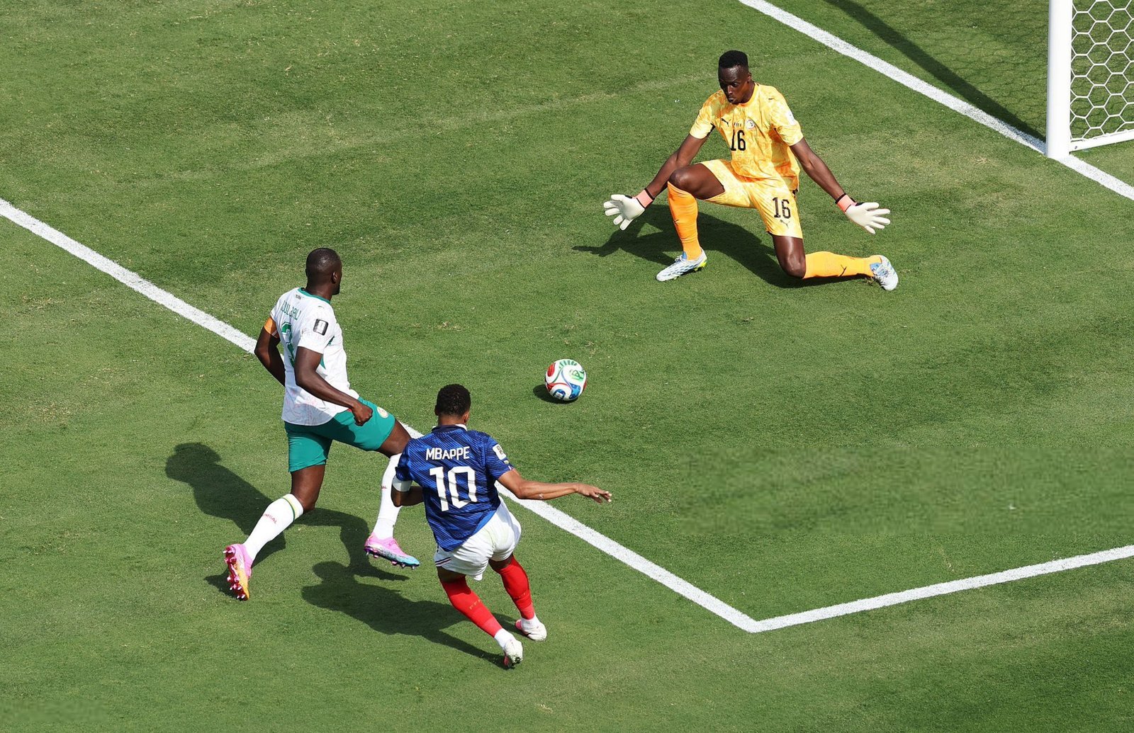 Overhead view of a soccer match: yellow-clad goalkeeper (number 16) dives near the goal as attackers close in on the ball while on a green pitch.