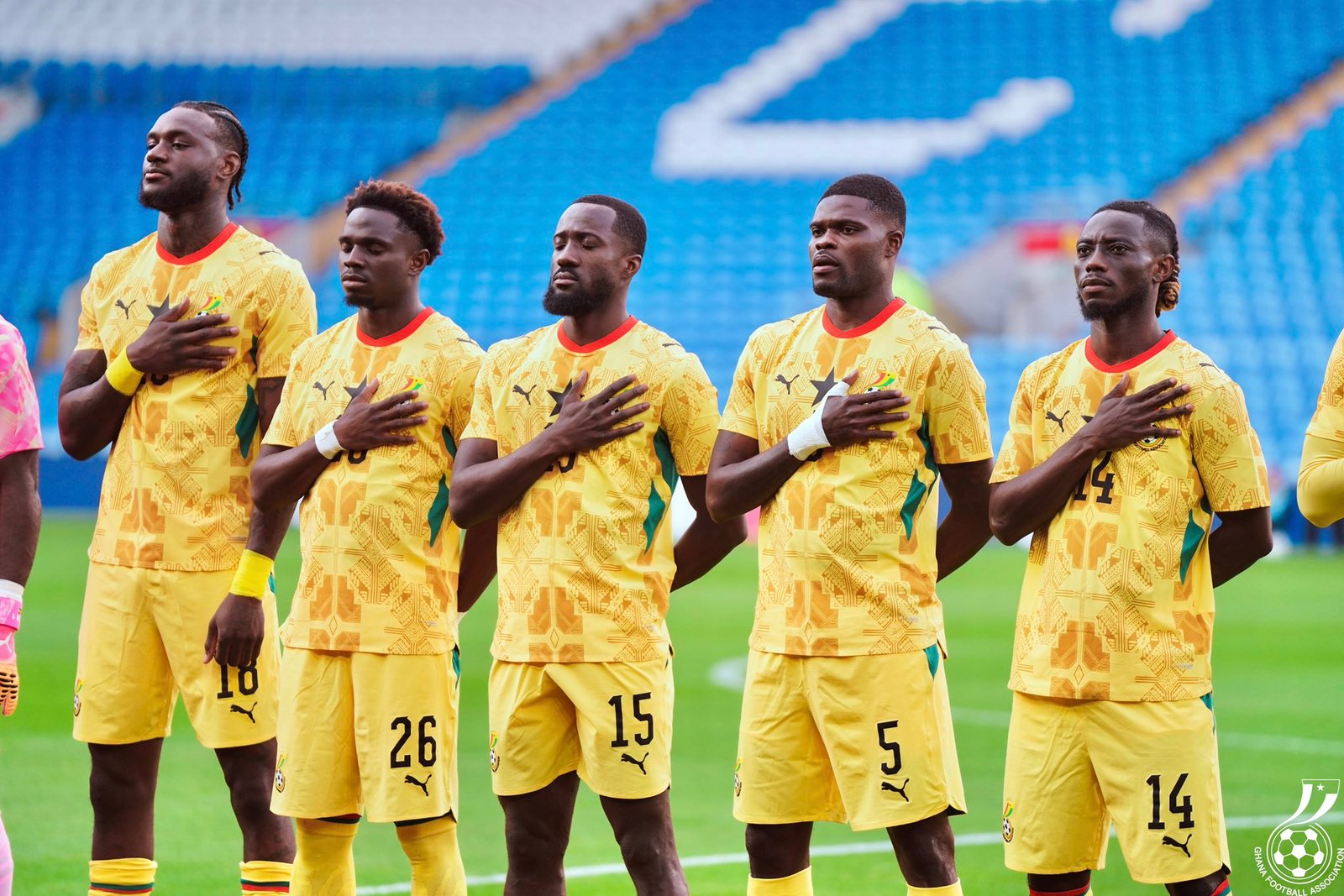 Group of Ghanaian footballers in yellow away kits standing in a line with hands over their chests on a green pitch as anthem plays. Group of Ghanaian footballers in yellow away kits standing in a line with hands over their chests on a green pitch as anthem plays.