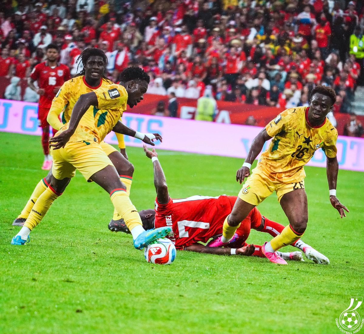 Soccer match action: two players in yellow jerseys challenge for the ball while a player in red lies on the ground on a green pitch, with a crowded stadium in the background. Soccer match action: two players in yellow jerseys challenge for the ball while a player in red lies on the ground on a green pitch, with a crowded stadium in the background.