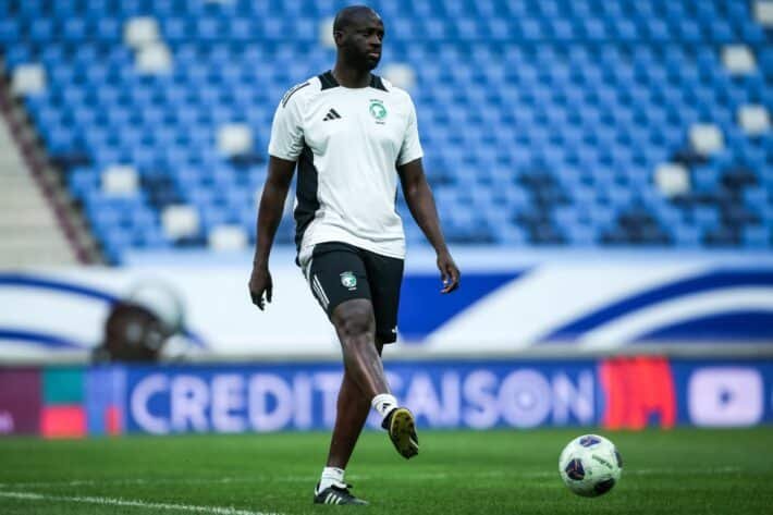 Soccer player in a white jersey dribbling a ball on a green pitch with blue stadium seating in the background Soccer player in a white jersey dribbling a ball on a green pitch with blue stadium seating in the background