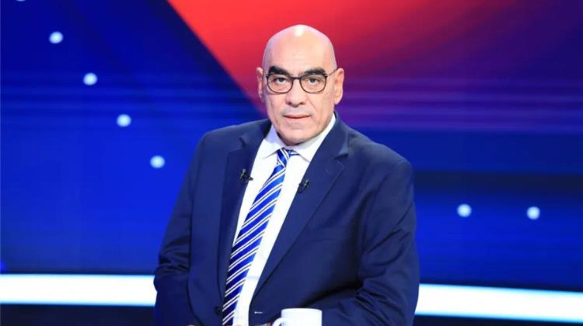 Male news anchor in a navy suit and striped tie sits in a blue-and-red television studio background with a thoughtful expression.