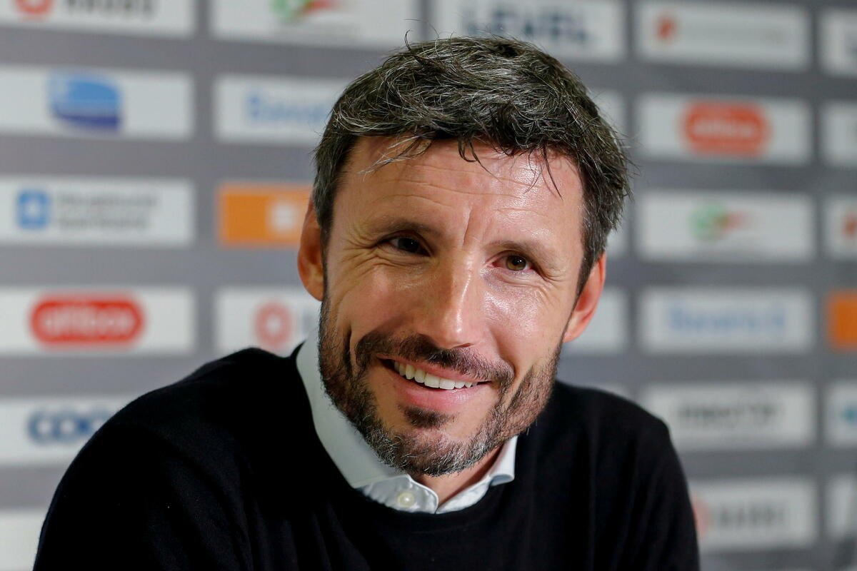 Smiling man with dark hair and stubble at a press conference, sponsor logos blurred in the background.