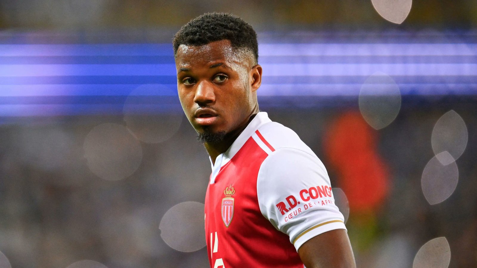 Soccer player in a red-and-white jersey looks over his shoulder on the field with a blurred crowd and blue lights in the background.