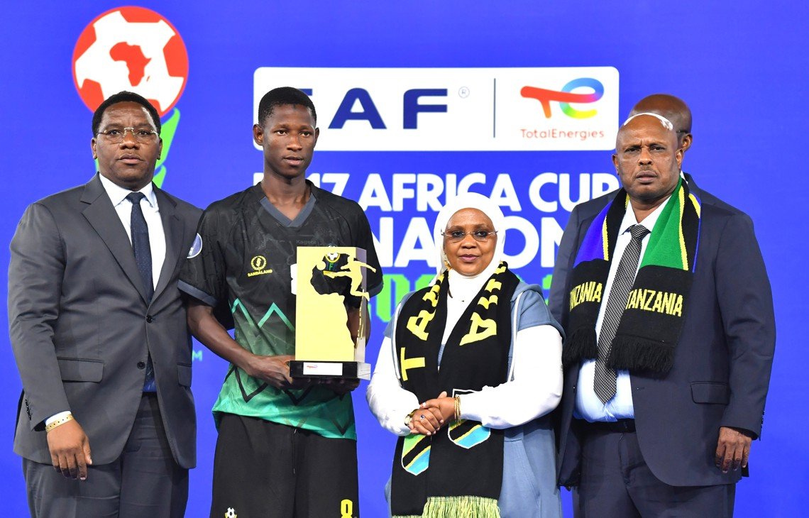 Athlete in black sportswear holding a trophy, flanked by two men in suits and a woman in a headscarf on a stage with a blue backdrop and event logos. Athlete in black sportswear holding a trophy, flanked by two men in suits and a woman in a headscarf on a stage with a blue backdrop and event logos. Athlete in black sportswear holding a trophy, flanked by two men in suits and a woman in a headscarf on a stage with a blue backdrop and event logos. Athlete in black sportswear holding a trophy, flanked by two men in suits and a woman in a headscarf on a stage with a blue backdrop and event logos.