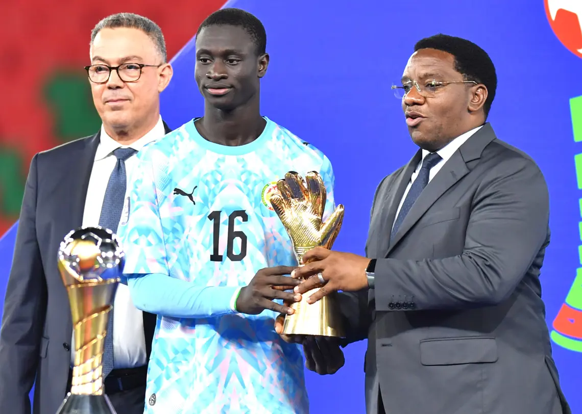 Young goalkeeper in a light blue jersey with number 16 accepts a trophy from a suited man at an award ceremony, with another man nearby holding a trophy. Young goalkeeper in a light blue jersey with number 16 accepts a trophy from a suited man at an award ceremony, with another man nearby holding a trophy. Young goalkeeper in a light blue jersey with number 16 accepts a trophy from a suited man at an award ceremony, with another man nearby holding a trophy. Young goalkeeper in a light blue jersey with number 16 accepts a trophy from a suited man at an award ceremony, with another man nearby holding a trophy.