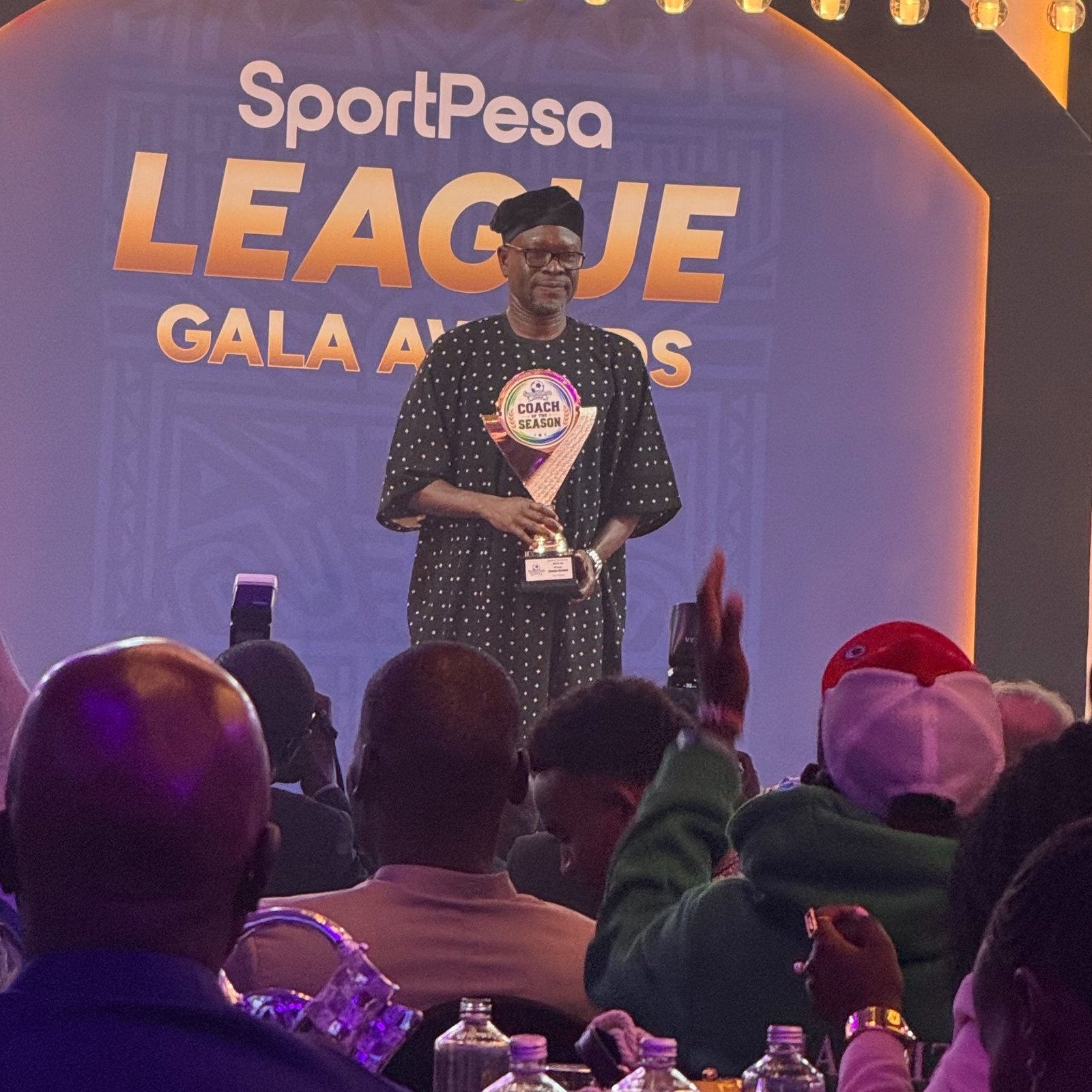 Man in traditional attire on stage holding a trophy at a SportPesa League Gala Awards event, with audience in foreground and a blue backdrop. Man in traditional attire on stage holding a trophy at a SportPesa League Gala Awards event, with audience in foreground and a blue backdrop.