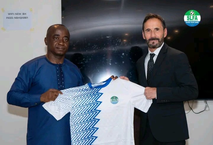 Two men in traditional blue attire and a suit hold up a white SLFA soccer jersey with a blue zigzag design.