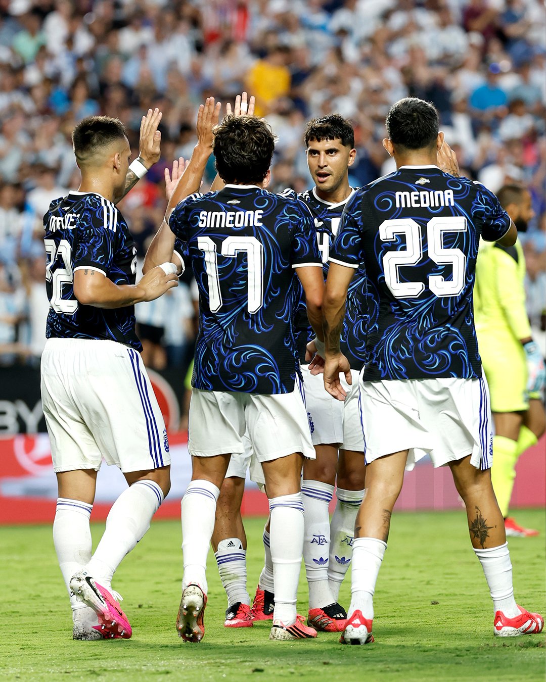 Soccer players in blue patterned jerseys celebrate on a green field, giving high-fives as a crowd watches.