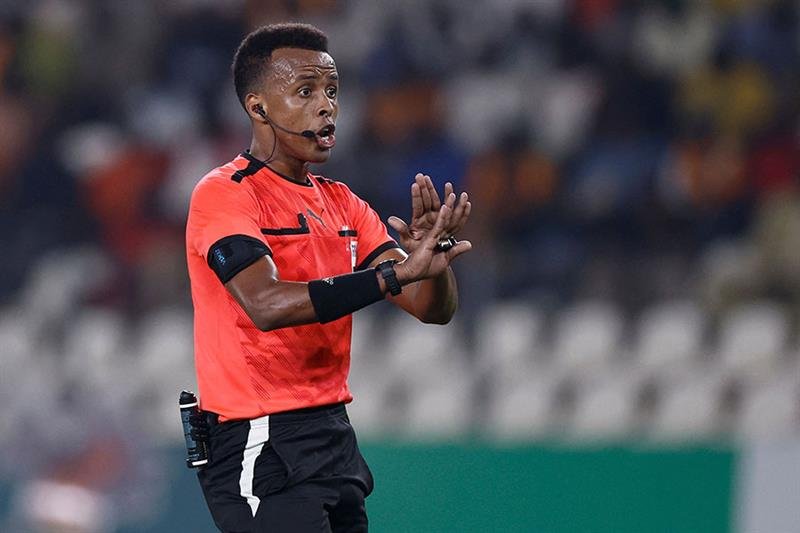Soccer referee in an orange jersey signaling with hands during a match, wearing a headset and a radio on his belt.