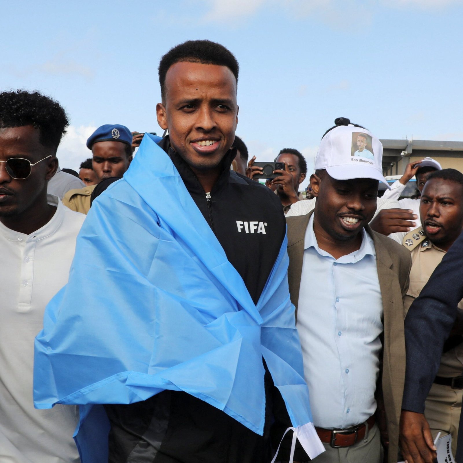Central man wearing a blue jacket with a FIFA logo smiles at a crowded outdoor event against a clear blue sky; several people surround him, some in uniforms and hats.