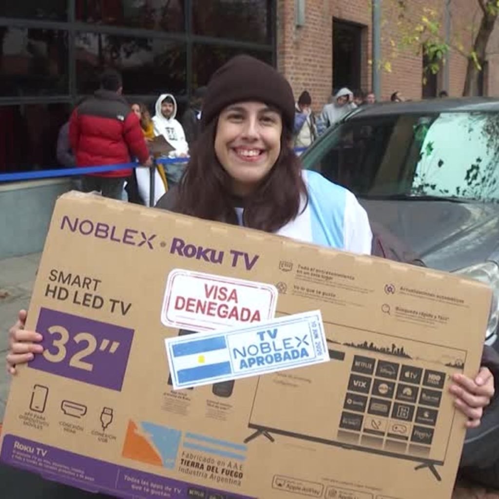 Smiling woman in a brown beanie holding a large 32-inch Roku TV box outdoors, with two stickers on the front.