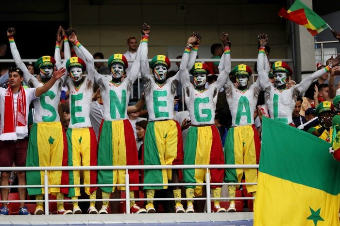 Group of Senegal fans with painted chests spelling 'SENEGAL', wearing colorful outfits at a stadium.