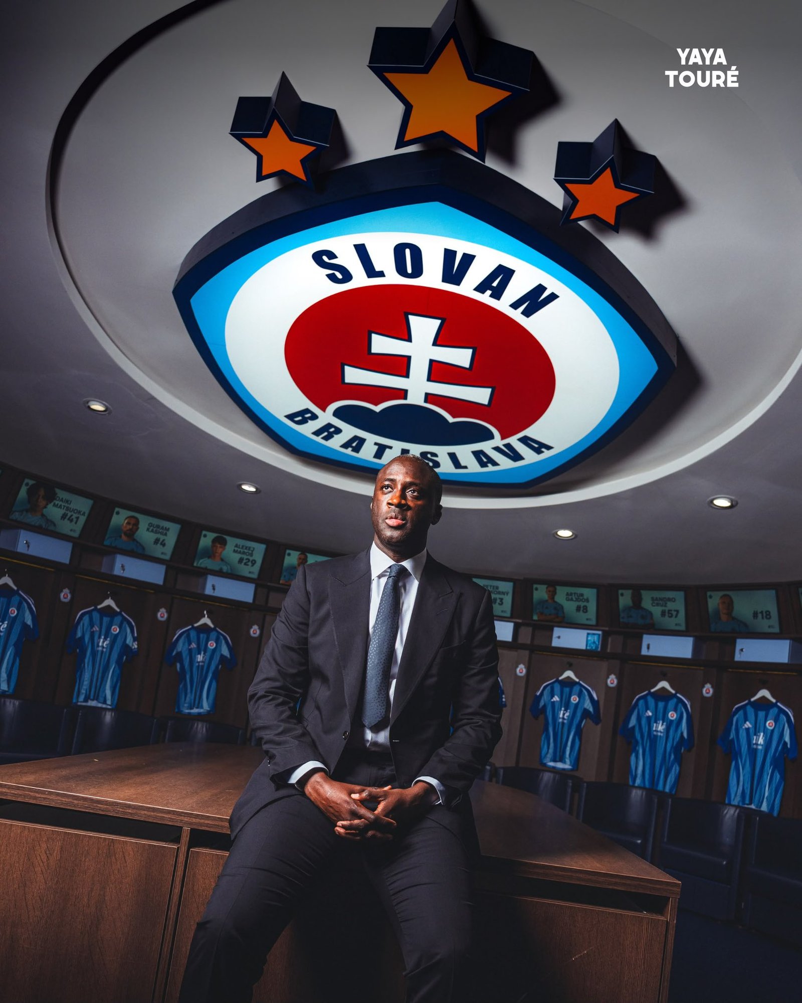 Man in a dark suit seated at a wooden table inside a soccer locker room, with the Slovakia crest overhead.