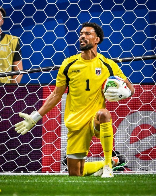 Goalkeeper in yellow jersey with number 1 kneels in front of the goal, holding a soccer ball and wearing white gloves.
