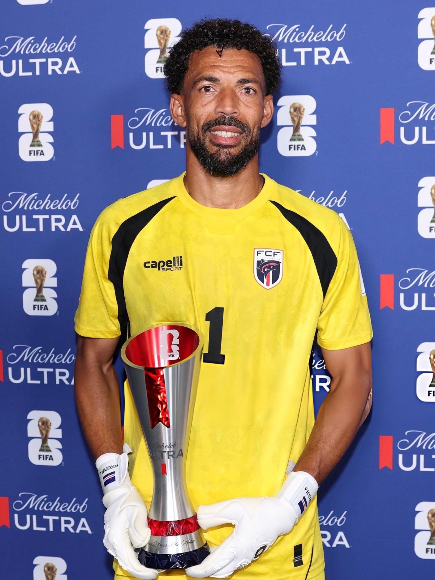 Male soccer goalkeeper in a yellow jersey holding a tall silver trophy, wearing white gloves, against a blue Michelob Ultra backdrop.
