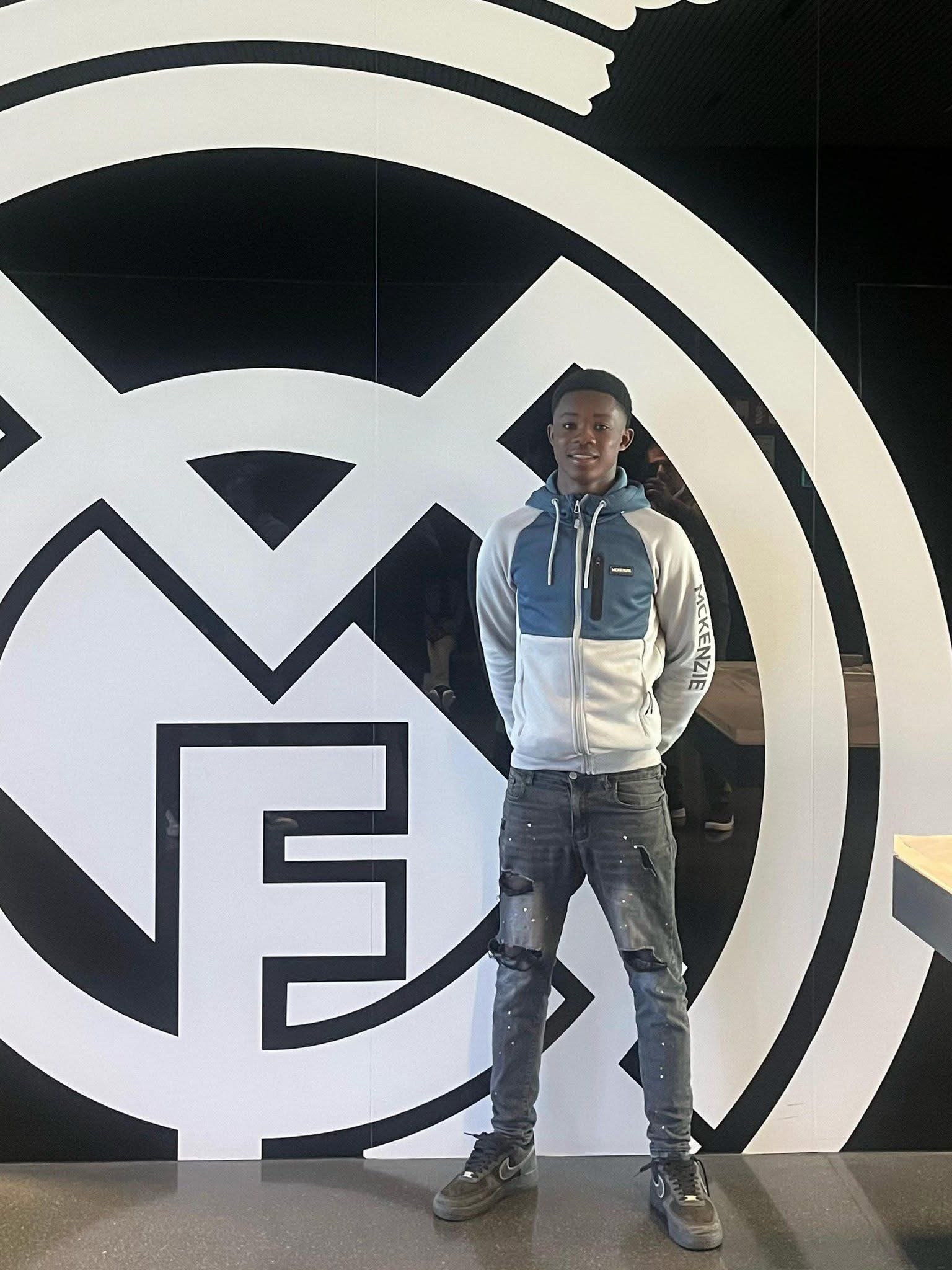 Young man in a blue-gray hoodie and distressed jeans posing in front of a large black-and-white circular mural backdrop.