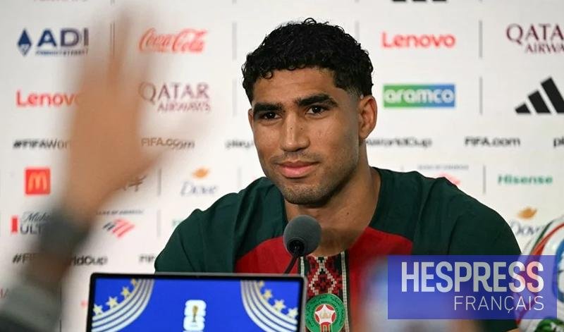 Man at a press conference speaking into a microphone, wearing a green and red shirt; blurred hand in the foreground and sponsor logos on a backdrop, with a blue 'HESPRESS FRANÇAIS' overlay. Man at a press conference speaking into a microphone, wearing a green and red shirt; blurred hand in the foreground and sponsor logos on a backdrop, with a blue 'HESPRESS FRANÇAIS' overlay.