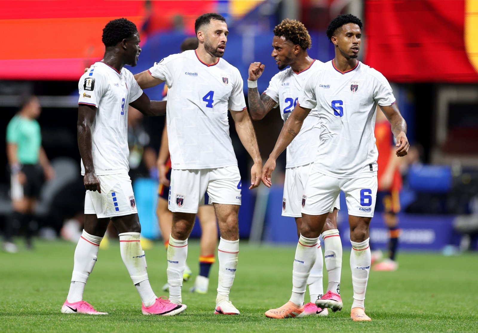 Four soccer players in white uniforms stand together on a green pitch, celebrating with fists and arms around each other.