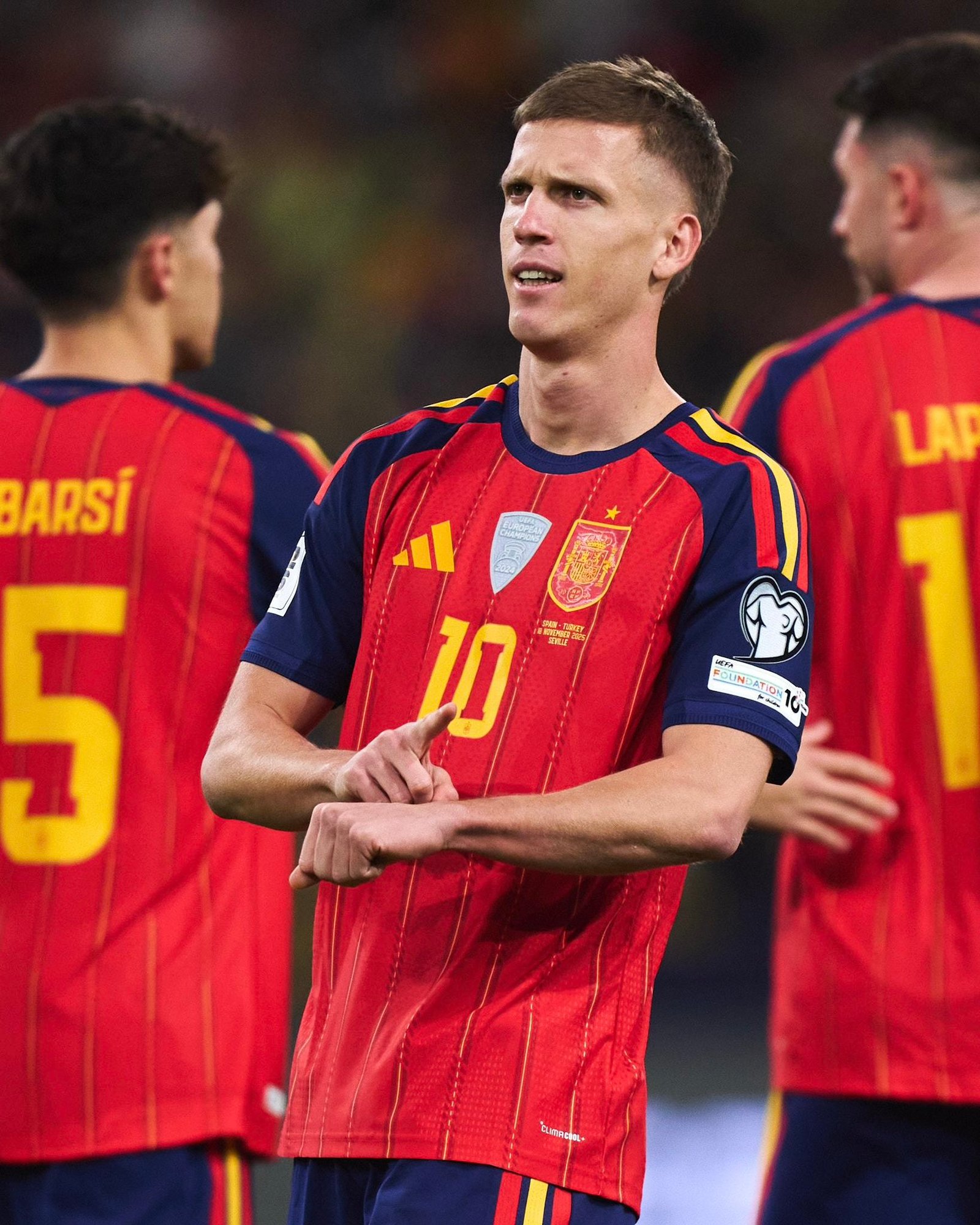 Centered player in red Spain kit (number 10) with blue shorts, looks ahead while others in red meet around him on the field during a celebration.
