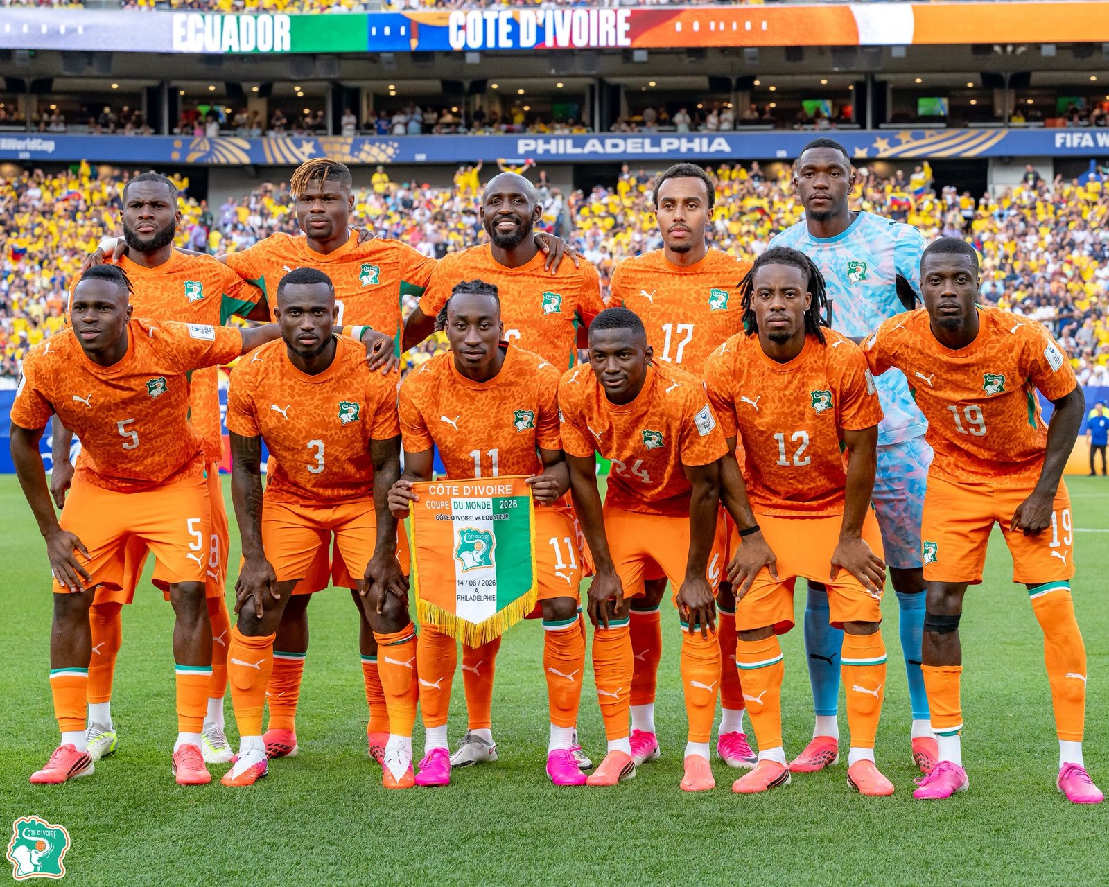 Ivory Coast national football team players in orange jerseys pose for a group photo on a green pitch with yellow-clad fans in the stands behind them.