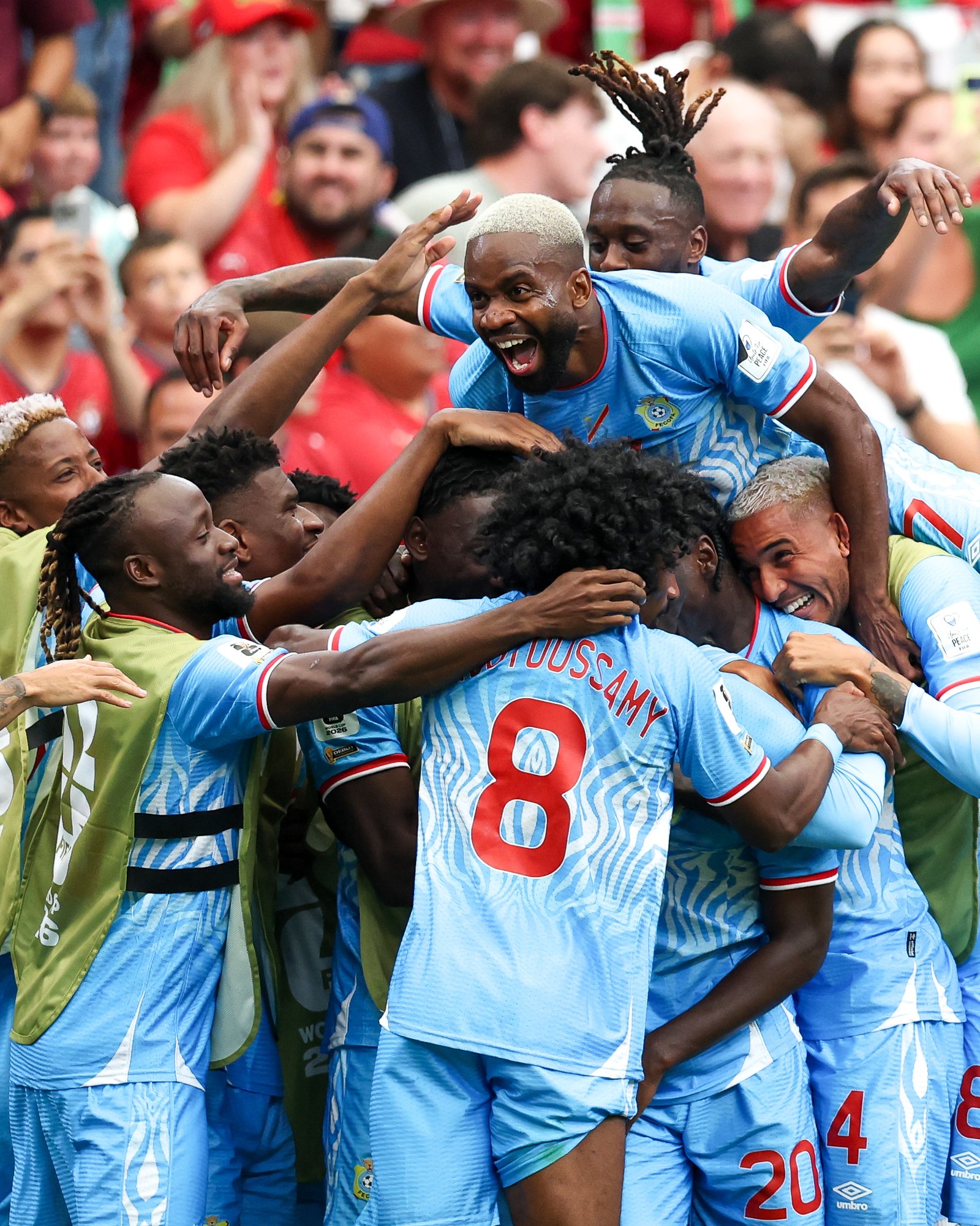 Soccer players in light blue uniforms celebrate in a crowded huddle, arms raised and smiling.