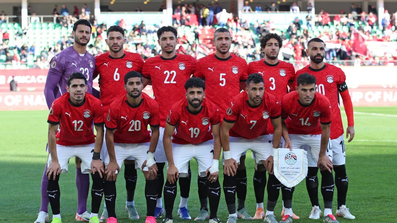 Egyptian football team in red jerseys posing for a group photo on the pitch with fans in the stands behind them. Egyptian football team in red jerseys posing for a group photo on the pitch with fans in the stands behind them.