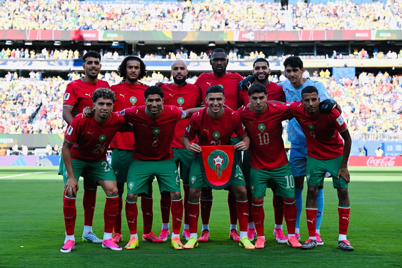 Moroccan national team players pose for a group photo on the pitch with a trophy in front and a packed stadium behind them. Moroccan national team players pose for a group photo on the pitch with a trophy in front and a packed stadium behind them.