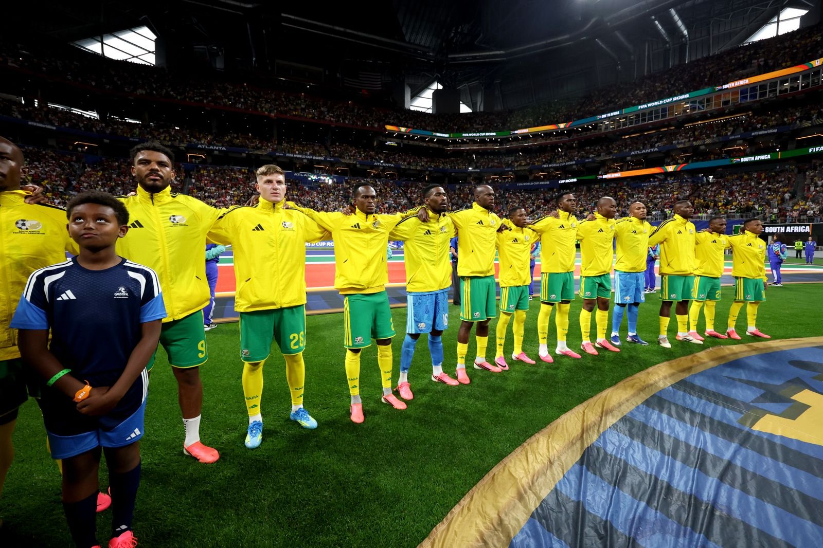 National soccer team players in yellow jerseys stand shoulder-to-shoulder on the field with a child in front, during a crowded stadium ceremony. National soccer team players in yellow jerseys stand shoulder-to-shoulder on the field with a child in front, during a crowded stadium ceremony.