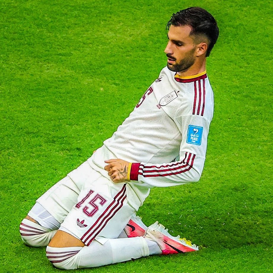 Soccer player kneeling on grass in a white kit with red trim, number 15, blue peace badge on the sleeve. Soccer player kneeling on grass in a white kit with red trim, number 15, blue peace badge on the sleeve.