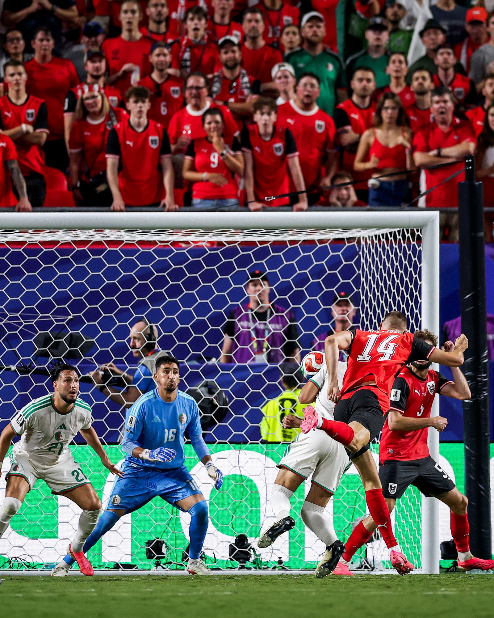 Soccer match scene: red-clad attackers press near the goal as a defender and goalkeeper in blue defend; crowd cheers in the stands behind the net. Soccer match scene: red-clad attackers press near the goal as a defender and goalkeeper in blue defend; crowd cheers in the stands behind the net.
