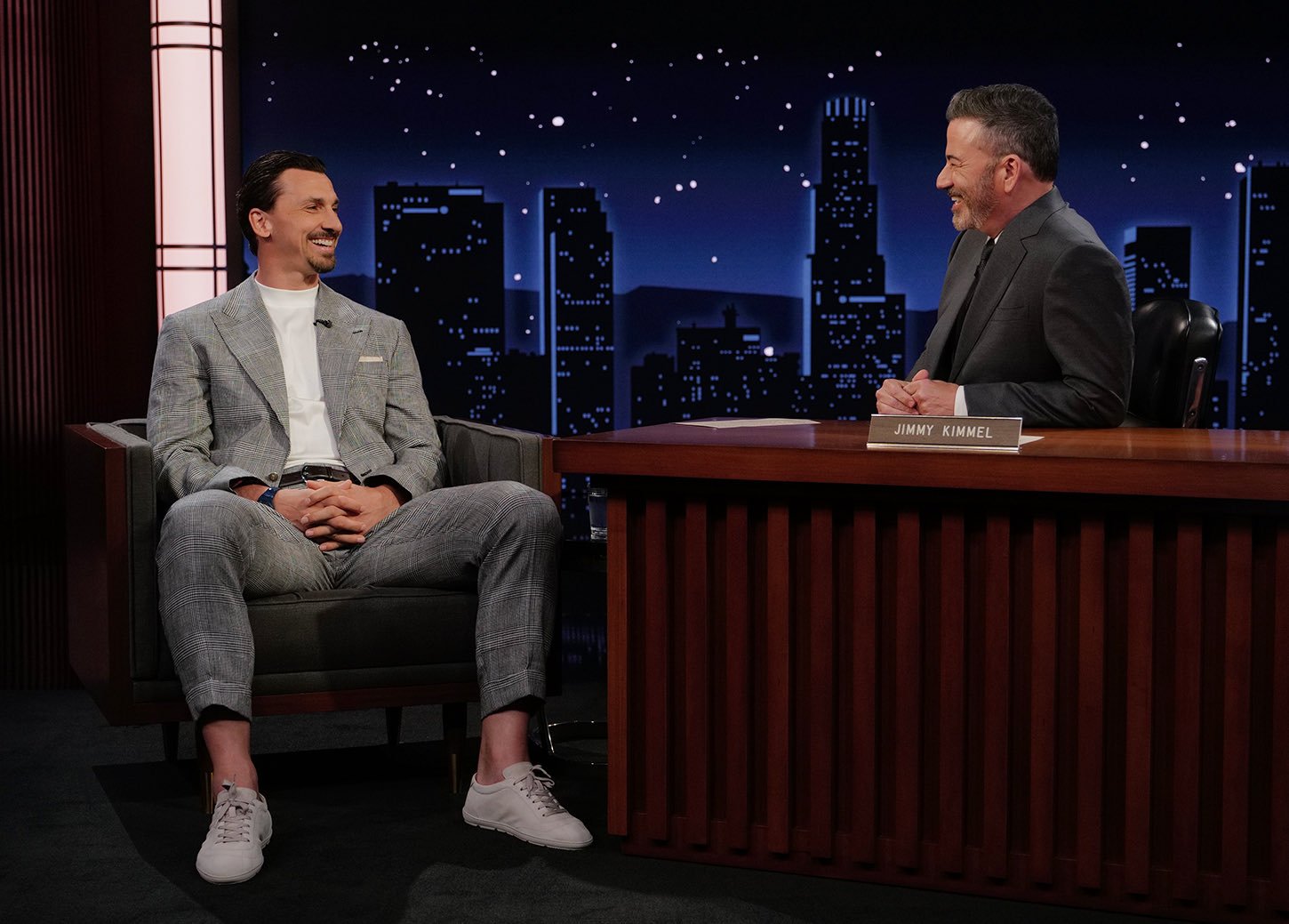 Talk show host interviewing a smiling guest in a light gray plaid suit on a studio set with a nighttime cityscape backdrop.