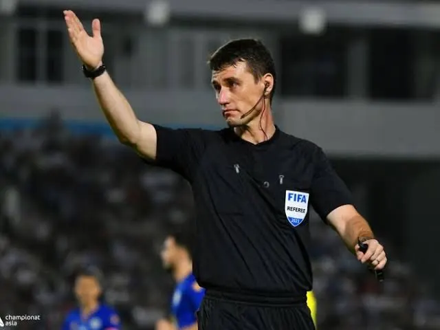 FIFA referee in black uniform raises left arm, signaling a call during a match in a stadium.