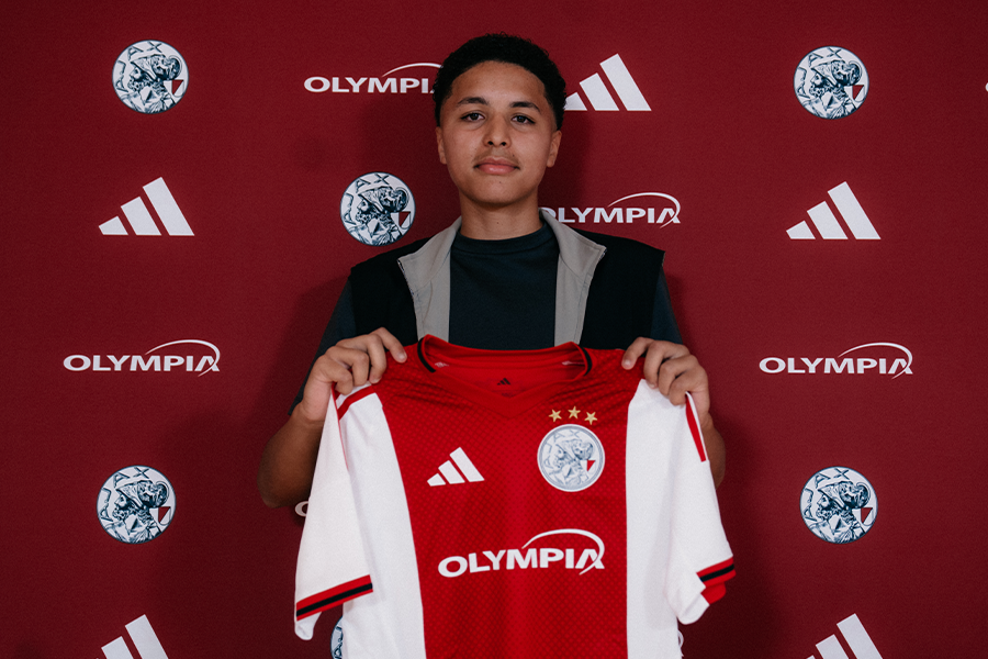 Person standing in front of a maroon backdrop with Olympia logos, holding a red and white Adidas soccer jersey with a crest and stars. Person standing in front of a maroon backdrop with Olympia logos, holding a red and white Adidas soccer jersey with a crest and stars.