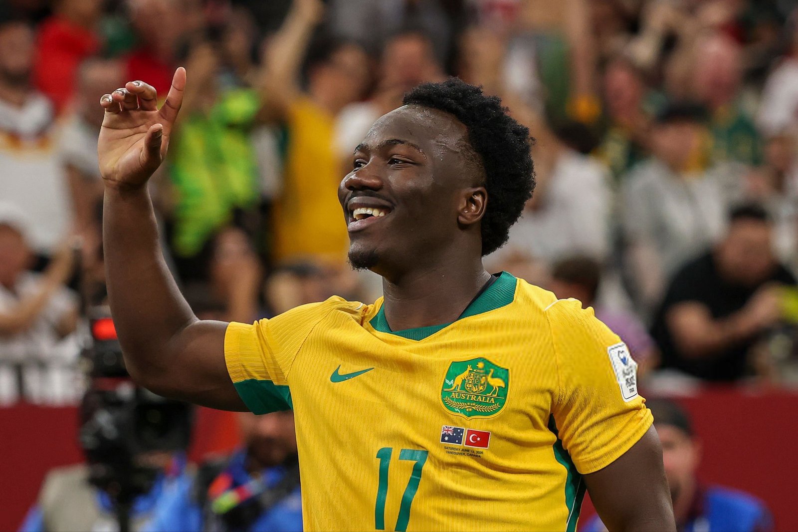 Australian soccer player in a yellow jersey with number 17 celebrates, arm raised, crowded stadium in the background.