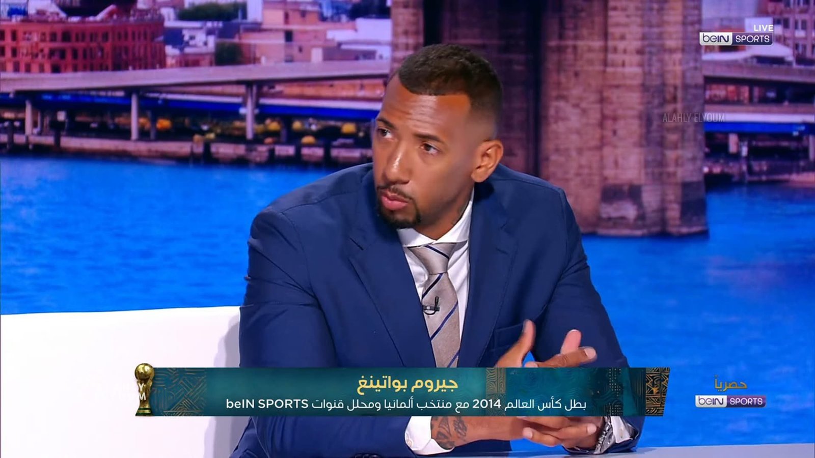 Male guest in a blue suit speaks on a TV talk show, with a cityscape backdrop and a green Arabic banner across the bottom.