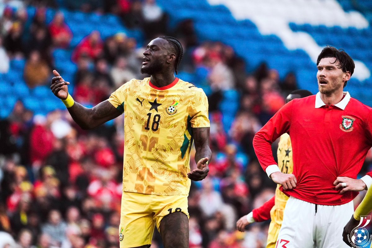 Ghanaian footballer in a yellow kit with number 18 gestures with his arm during a match, while a Welsh opponent in red stands nearby in a stadium filled with spectators. Ghanaian footballer in a yellow kit with number 18 gestures with his arm during a match, while a Welsh opponent in red stands nearby in a stadium filled with spectators.