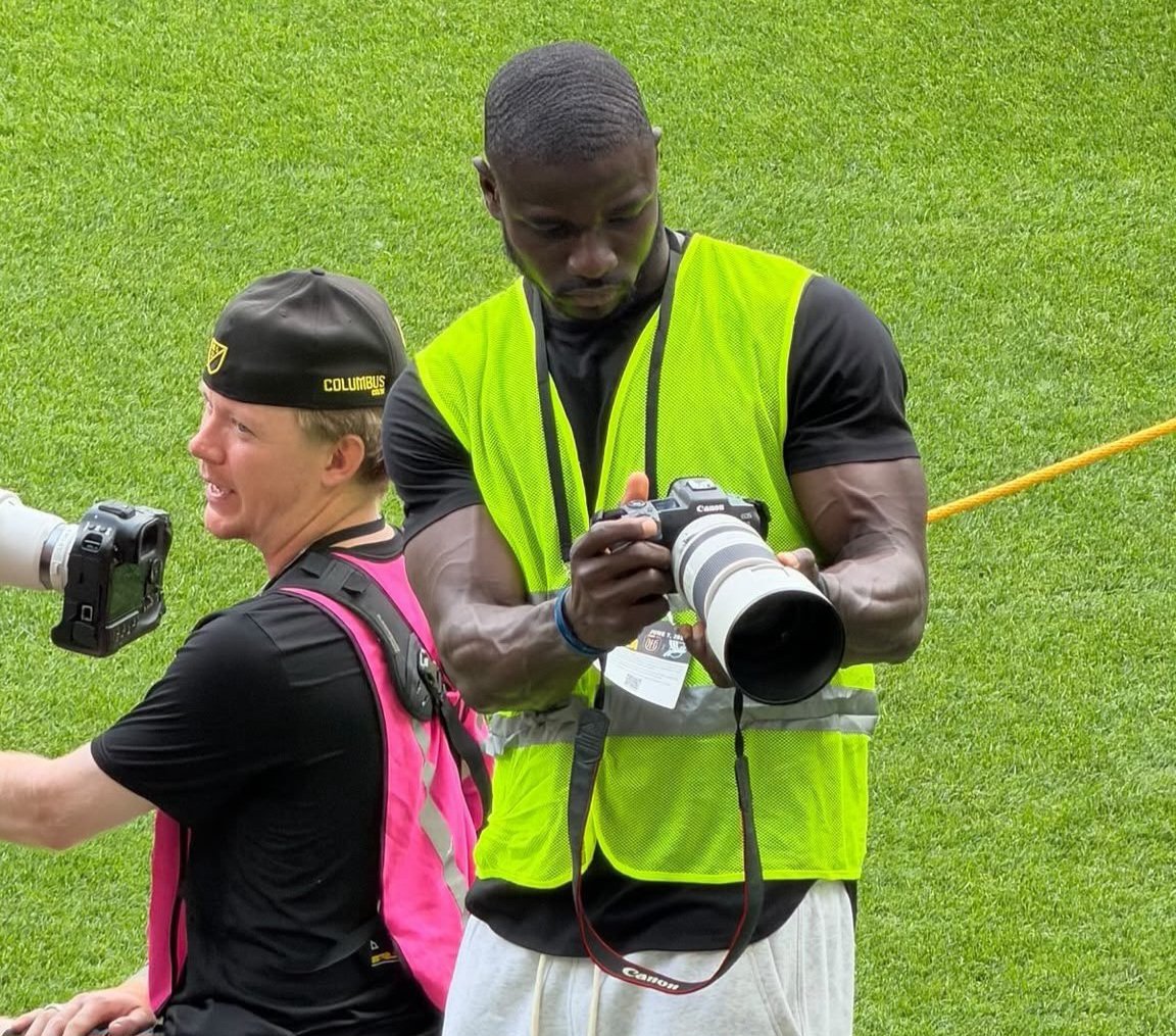 Two photographers kneel on a grassy field, one in a bright yellow safety vest with a large telephoto lens ready to shoot as the other wears a pink vest and black cap.
