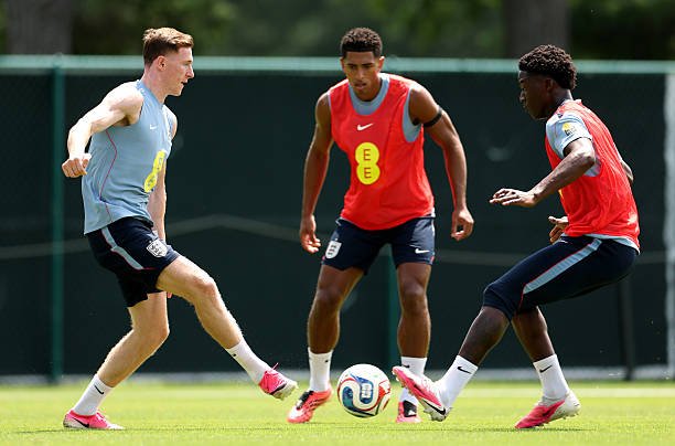 Three soccer players in practice bibs contest a ball on a sunny field, one preparing to kick as two defend nearby. Three soccer players in practice bibs contest a ball on a sunny field, one preparing to kick as two defend nearby.