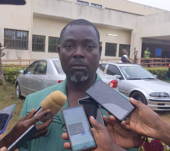 Man giving a press interview outside a beige building, with microphones and smartphones held up to him by reporters. Man giving a press interview outside a beige building, with microphones and smartphones held up to him by reporters.