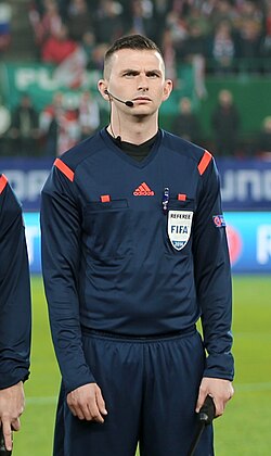 Football referee in a navy kit with a FIFA badge and headset standing on a football pitch. Football referee in a navy kit with a FIFA badge and headset standing on a football pitch.