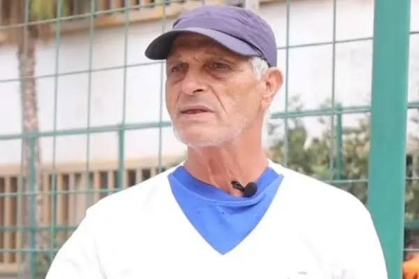 Older man wearing a purple cap and white shirt with a blue undershirt, standing near a green chain-link fence outdoors. Older man wearing a purple cap and white shirt with a blue undershirt, standing near a green chain-link fence outdoors.