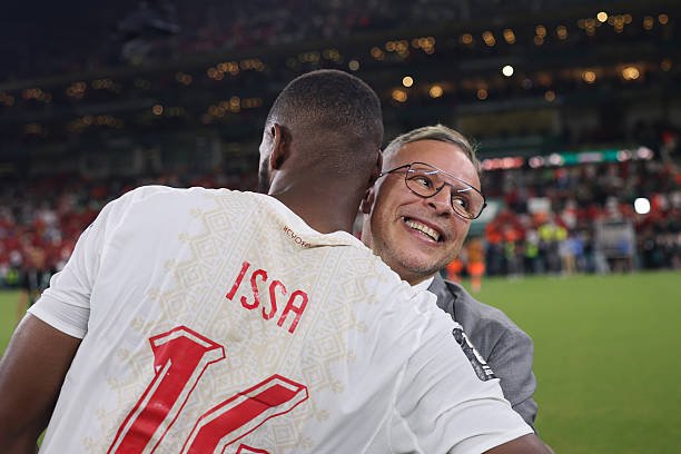 Soccer player wearing white jersey with ISSA and number 12 hugs a smiling man in glasses on a field after a match in a stadium. Soccer player wearing white jersey with ISSA and number 12 hugs a smiling man in glasses on a field after a match in a stadium.