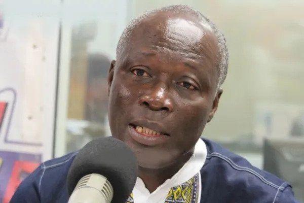 Man being interviewed on a microphone in a studio, speaking with a focused expression and wearing a dark jacket over a white collared shirt.