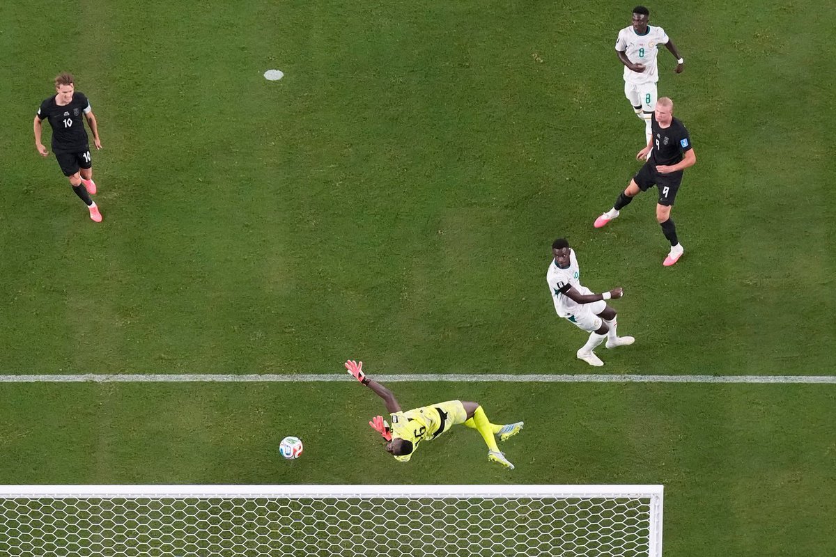 A goalkeeper in yellow dives to save a low ball near the goal line as players in black and white chase nearby on a green pitch.