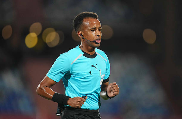 Soccer referee in a bright blue uniform with headset, running during a match with a focused expression.