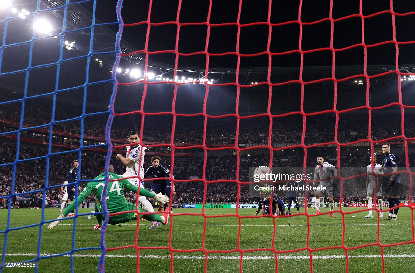Soccer scene from the goal area: goalkeeper in green dives to block a shot as players contest near the goal with a full stadium behind. Soccer scene from the goal area: goalkeeper in green dives to block a shot as players contest near the goal with a full stadium behind.