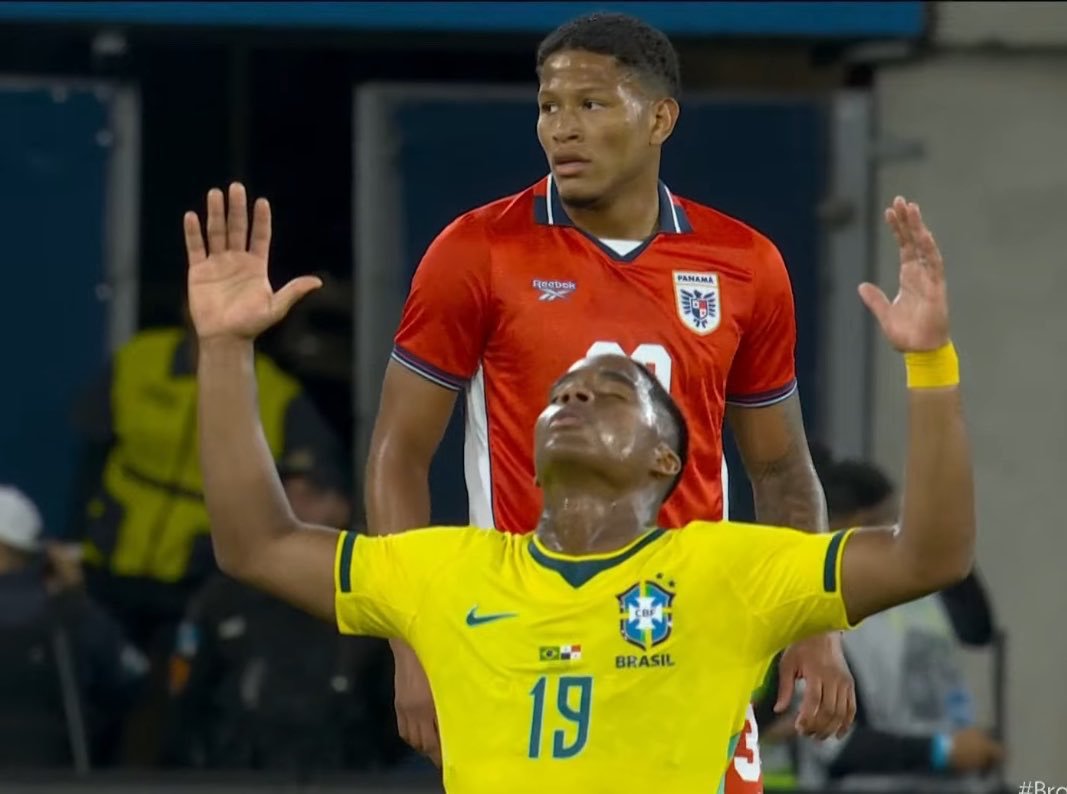Brazil player in yellow raises arms in celebration, eyes closed, with a red-jersey opponent standing behind him.