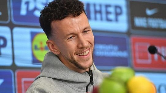 Smiling man in a gray hoodie at a press conference, microphone in front, colorful sponsor banners blurred in the background. Smiling man in a gray hoodie at a press conference, microphone in front, colorful sponsor banners blurred in the background.