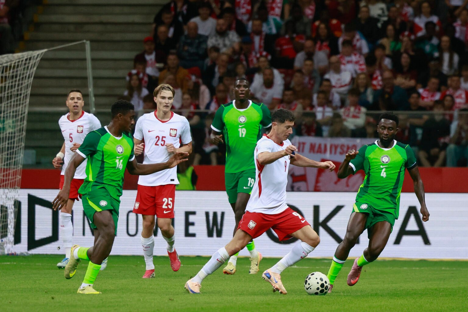 Soccer match with players in white and red versus green kits battling for the ball near the goal area, crowded stands in the background
