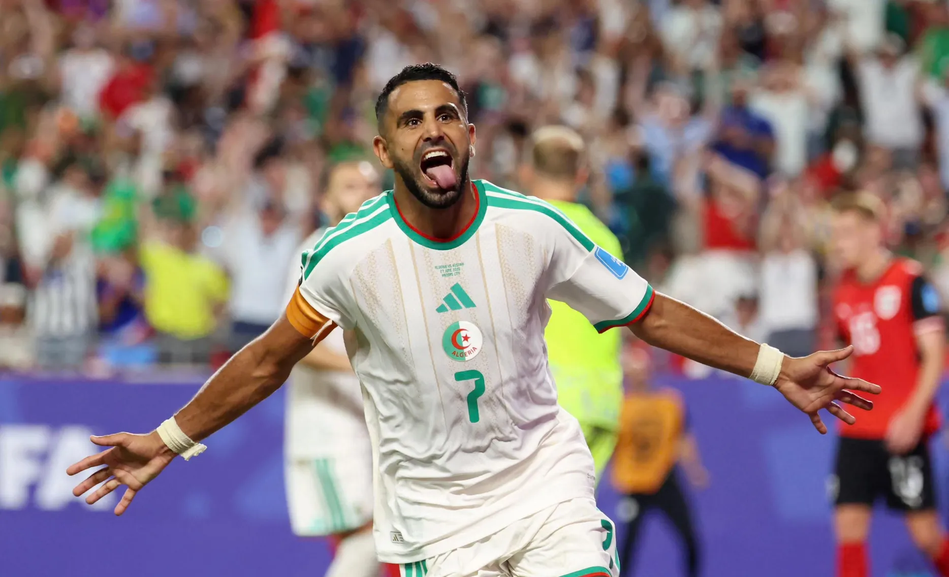 Algerian soccer player celebrating with arms outstretched and tongue out on a crowded stadium field Algerian soccer player celebrating with arms outstretched and tongue out on a crowded stadium field